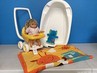 Lot containing a yellow Little Tikes toy stroller, doll, blue stuffed animal, colorful blanket, and white baby bathtub against a blue background.