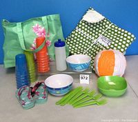Photo of full picnic set including green floral tote bag, stacked colorful plastic cups, water bottle, white and blue bowls, green bowl, lime green forks, beach ball, and children's flip flops.