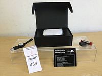 Black box containing the power box device and accessories on clear display stand with auction tag