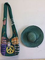 Large green fabric shoulder bag with colorful fringe and peace symbol embroidery next to green woven wicker sun hat.