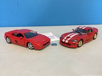 Front and side views of two red diecast model cars, one a Ferrari and the other a Dodge Viper with racing stripes, placed on a grey surface against a blue background.