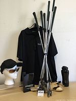 Photo displaying vintage golf clubs, black golf shirt hanging on wall, sunglasses on head mannequin worn with black cap, and items including vintage golf shoes and water bottle on table.