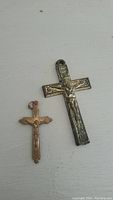 Photo showing two crucifix pendants, one larger with ornate detailing and one smaller copper-toned, both depicting Christ