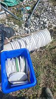 Photo shows a pile of large white plastic buckets stacked horizontally on gravel with a blue tub filled with white bucket lids.