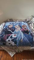 View of Frozen themed blanket with Queen Elsa, Princess Anna, Olaf and Kristoff spread flat on a bed.