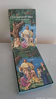 The Rainbow Bible standing upright with its matching protective box behind it, both showing colorful illustrated covers featuring Jesus and children.