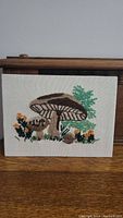 Full view of embroidered mushroom crewel art on fabric showing two large brown mushrooms with green leaves and small orange-yellow flowers.