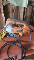 Side view of Ridgid electric jigsaw showing orange and grey casing, black handle grip and power cord coiled on surface.