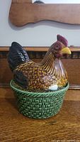 Rooster cookie jar with rooster lid on basket base shown from the side and front angle on wooden table.