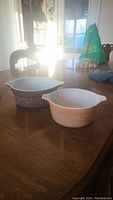 Two Corningware baking dishes on a wooden table, a square and a round dish with white interiors and brown woodland floral pattern exteriors.
