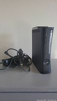 Black Xbox 360 Elite console standing vertically with power and AV cables beside it, showing front and side vents and buttons.