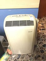 Full view of white DeLonghi portable air conditioner with front vent grille and attached hoses in background.