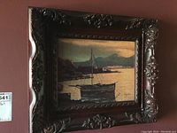 Framed painting showing a boat in water near rocky shoreline with mountains and buildings in the background