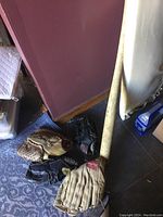 Photo of a wooden baseball bat and four baseball gloves arranged on the floor against wall, showing wear and usage.