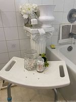 White ceramic column pedestal with faux flower arrangement, glass jar, frog dish, and white plastic bath seat in bathroom setting