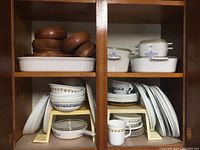 Shelved assortment of CorningWare casserole dishes with blue cornflower pattern, white serving dishes, plates, bowls, and wooden salad bowls and utensils.