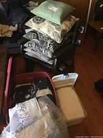 Photo of six throw pillows stacked on a chair with patterned and green covers, red plastic bin and white plastic bin on the floor next to the chair containing bedding and lace tablecloths