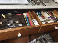 Photo showing three wooden drawers open with assorted cutlery and kitchen utensils organized inside, including spoons, forks, knives, peelers, can opener, and scissors.