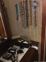 Photo showing numerous colorful beaded necklaces hanging on a wall-mounted jewelry holder above a dresser with watches and other small jewelry items.