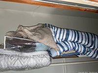 Side view of folded linens including patterned quilt and blue/white striped comforter stacked with rugs or curtains