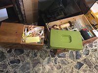 Three containers including a small wooden box, a green metal box, and a wooden drawer box on a stone floor containing sewing accessories.