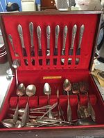 Open box showing an incomplete set of silver plated cutlery including knives in slots and various mixed pieces below in red velvet case