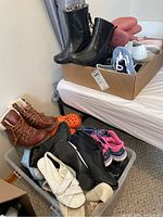 Photo showing multiple pairs of women's shoes including boots, sandals, and sneakers arranged in a corner and a cardboard box. Various materials and colors are visible including brown leather boots and black boots.