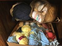 Top down view showing assortment of yarns stored in a cardboard box and plastic bag containing skeins in various colors including yellow, blue, red, and cream.