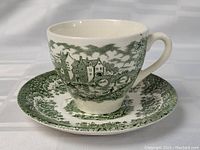 Tea cup and saucer set showing green transferware design of English village scene with houses and bridge on tea cup, matching floral and landscape border on saucer.
