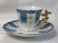 Photo showing tea cup and saucer set with intricate blue and gold hand painted design and ornate gold handle on white porcelain.