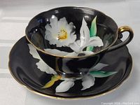 Tea cup and saucer set showing black porcelain with white floral decoration and gold trim.