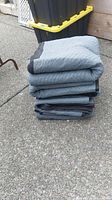 Stack of 5 gray moving blankets folded and stacked on pavement, showing quilted pattern and reinforced edges.