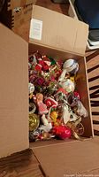 Open cardboard box filled with assorted vintage Christmas decorations including figurines and baubles in various colors and styles.