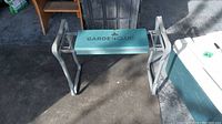 Front view of garden stool showing teal padded seat with 'Garden Club' branding and foldable metal frame.