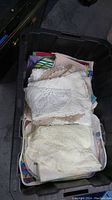 Photo showing a plastic tote filled with folded kitchen linens including crochet and lace tablecloths.