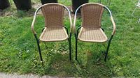 Front view of two lawn chairs placed on green grass. Chair frames are green tubular metal with woven tan wicker seats and backs.
