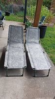 Two gray floral mesh fabric outdoor lounge chairs with green metal frames arranged side by side on concrete patio, with garden background.