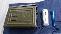 Metal military ammunition box with writing CAL .30 M1 AMMUNITION BOX, blue Maglite flashlight, and silver Garrity flashlight on blue cloth.