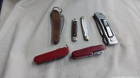 Full lot view showing six cutting tools arranged flat, including wooden handle knife, two small knives, two Swiss Army style red knives, and a large metallic folding knife with clip.