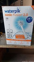 Front of retail box showing Waterpik Sonic-Fusion 2.0 branding and promotional details including up to 2x bristle tip speed and ADA acceptance.