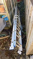 Two aluminum ladder sections stacked and stored outdoors beside a greenhouse and stone wall.