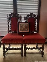 Pair of antique solid wood chairs with heavily carved backs and legs, upholstered in red fabric, shown with a decorative pillow placed on one seat.