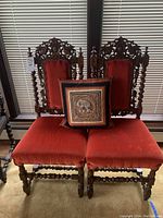 Front view of two antique wood chairs with carved backs and red upholstery, alongside a decorative pillow placed on seat