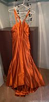 Orange satin gown front view hanging on white hanger indoors.