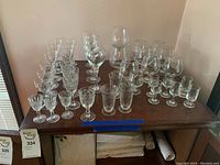 Top-down view of 39 assorted glass and crystal cups neatly arranged on a wooden table showing variety in glass styles.
