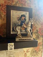 Figurine decanter of Marquis De Lafayette posed on a horse on a shelf with framed picture behind it.