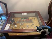 Wide photo showing miniature figurines arranged inside a glass display case on a table surface. Includes small decorative animals and a wooden cart miniature.