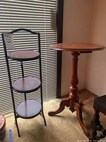 Two wooden side tables including a round wooden pedestal table and a black metal frame 3-tier wooden shelf, photographed against window blinds.