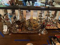 Wide view showing the full assortment of silver plated serving trays, candle sticks, and the silver polish canister on a wooden table.