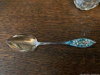 Close-up of teaspoon showing tarnished sterling surface and floral blue-green enamel handle.